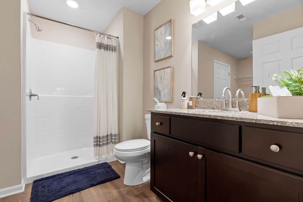 Modern bathroom with a glass-enclosed shower, toilet, dark wood vanity with granite countertop, mirror, wall art, and light fixtures; dark blue rug on wooden floor.