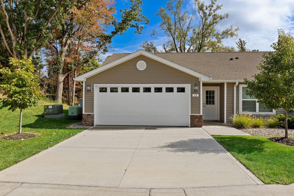 Single-story beige house with a two-car garage, concrete driveway, and a small lawn, surrounded by trees and utility boxes on the left.