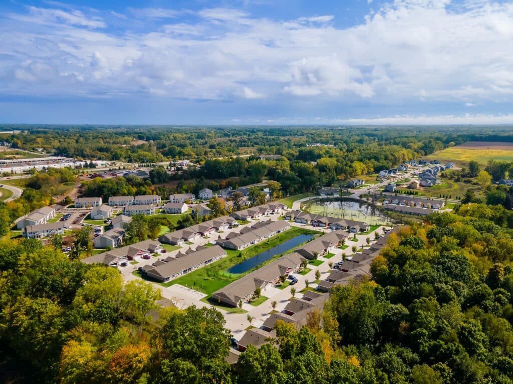 Aerial view of a suburban neighborhood with residential houses, streets, green spaces, ponds, and surrounding wooded areas under a partly cloudy sky.