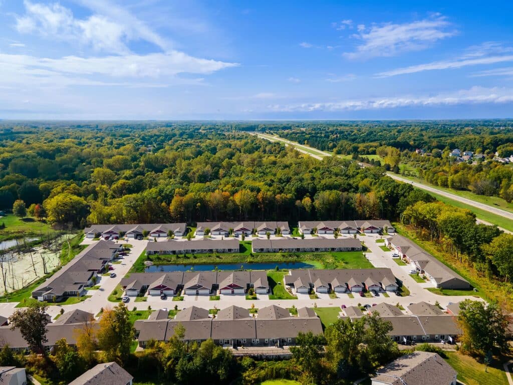 Aerial view of a suburban residential neighborhood with rows of houses, surrounded by trees and a highway in the background under a blue sky.