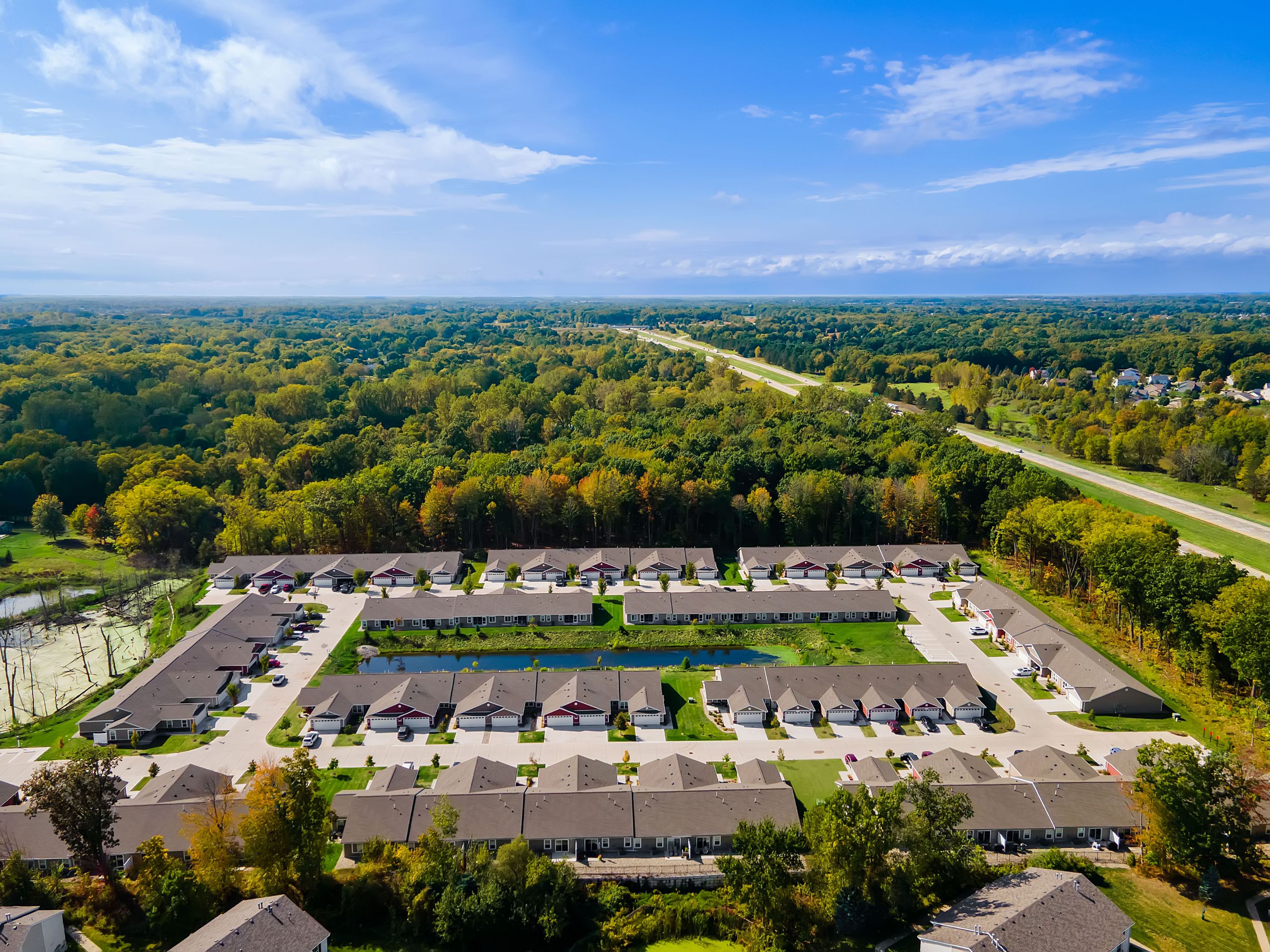 Aerial view of a suburban residential neighborhood with rows of houses, surrounded by trees and a highway in the background under a blue sky.