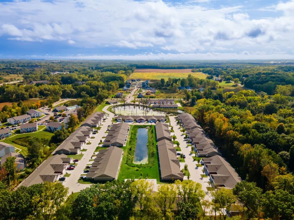 Aerial view of a suburban neighborhood with rows of houses surrounding a central pond, driveways, and trees, set amid green fields and forests under a partly cloudy sky.