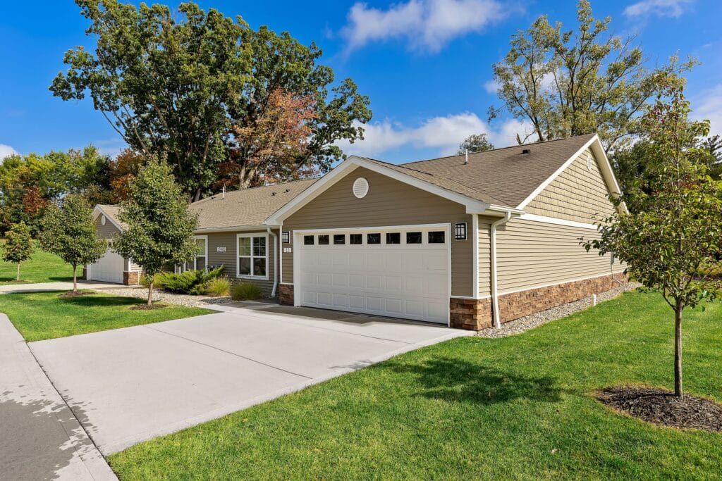 Single-story house with beige siding, a two-car garage, driveway, neatly trimmed lawn, and young trees, set in a suburban neighborhood with blue sky and leafy trees in the background.