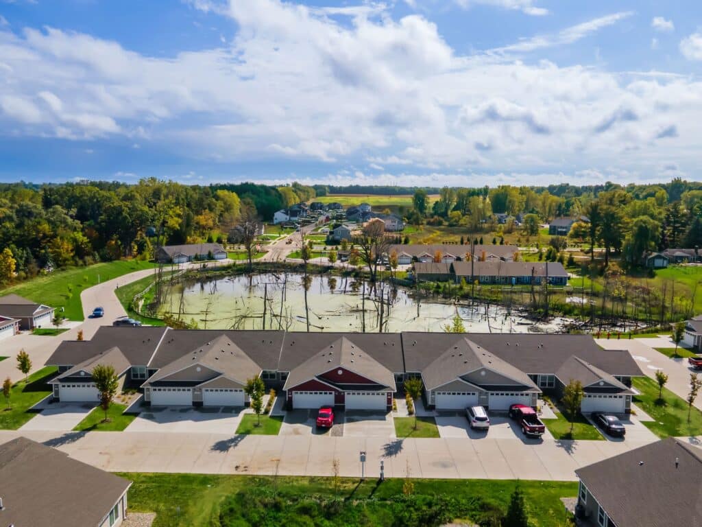A row of suburban houses with driveways and parked cars faces a small pond with trees, set in a green, residential neighborhood under a partly cloudy sky.