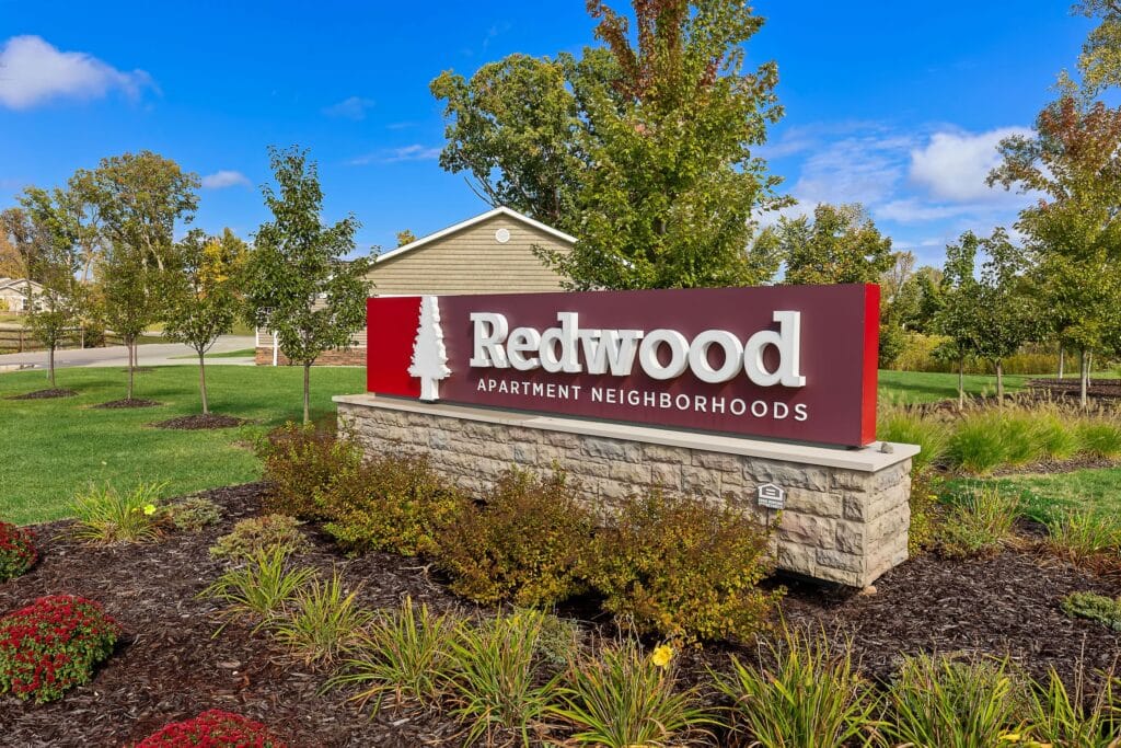 A stone and red sign reading "Redwood Apartment Neighborhoods" stands in landscaped grounds with green grass, shrubs, trees, and a clear blue sky.