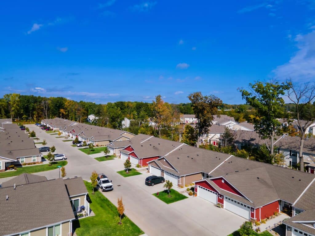 Aerial view of a suburban neighborhood with single-story houses, neatly arranged along a wide street under a clear blue sky. Cars are parked in driveways and trees are scattered throughout.