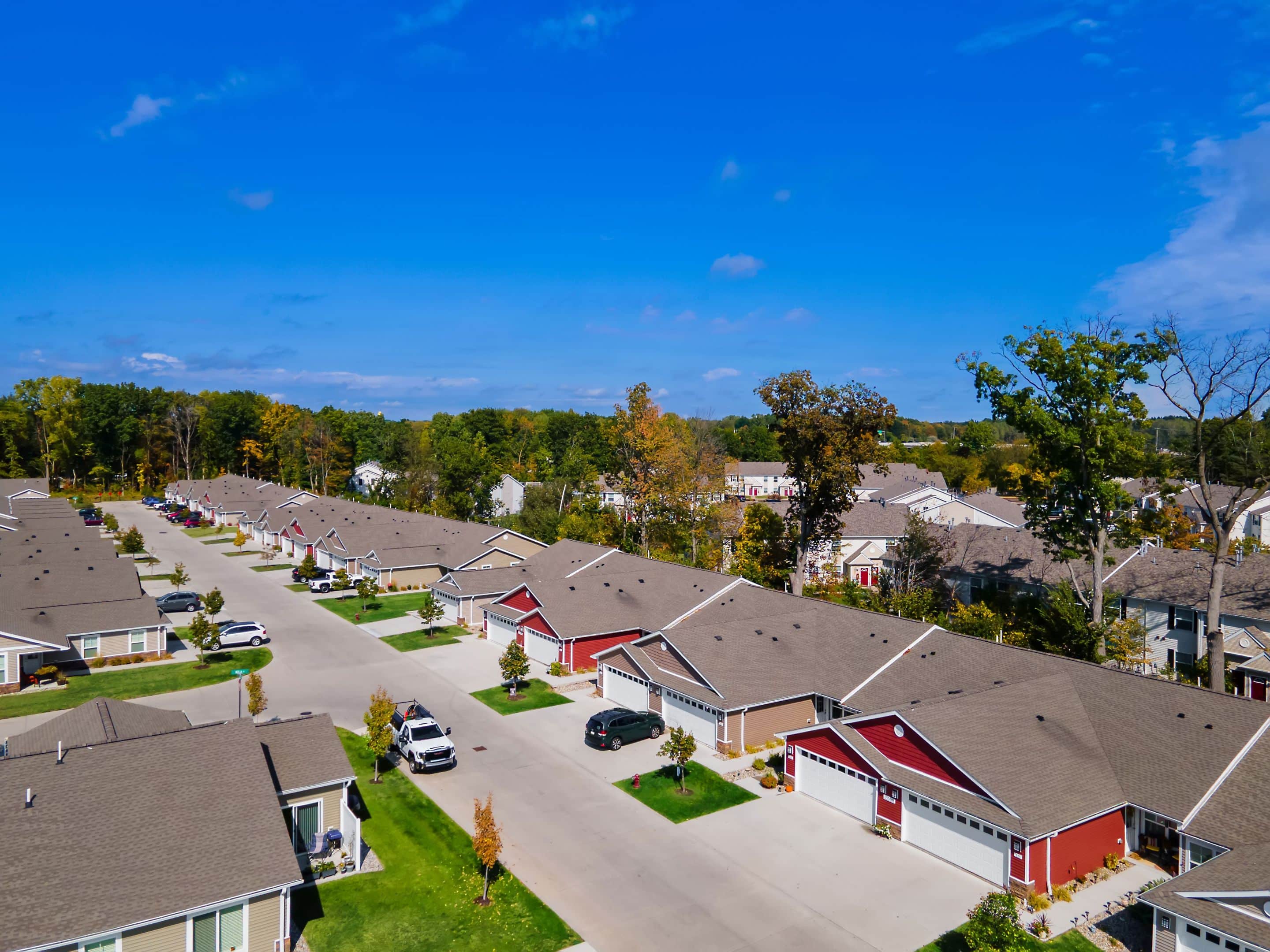Aerial view of a suburban neighborhood with single-story houses, neatly arranged along a wide street under a clear blue sky. Cars are parked in driveways and trees are scattered throughout.