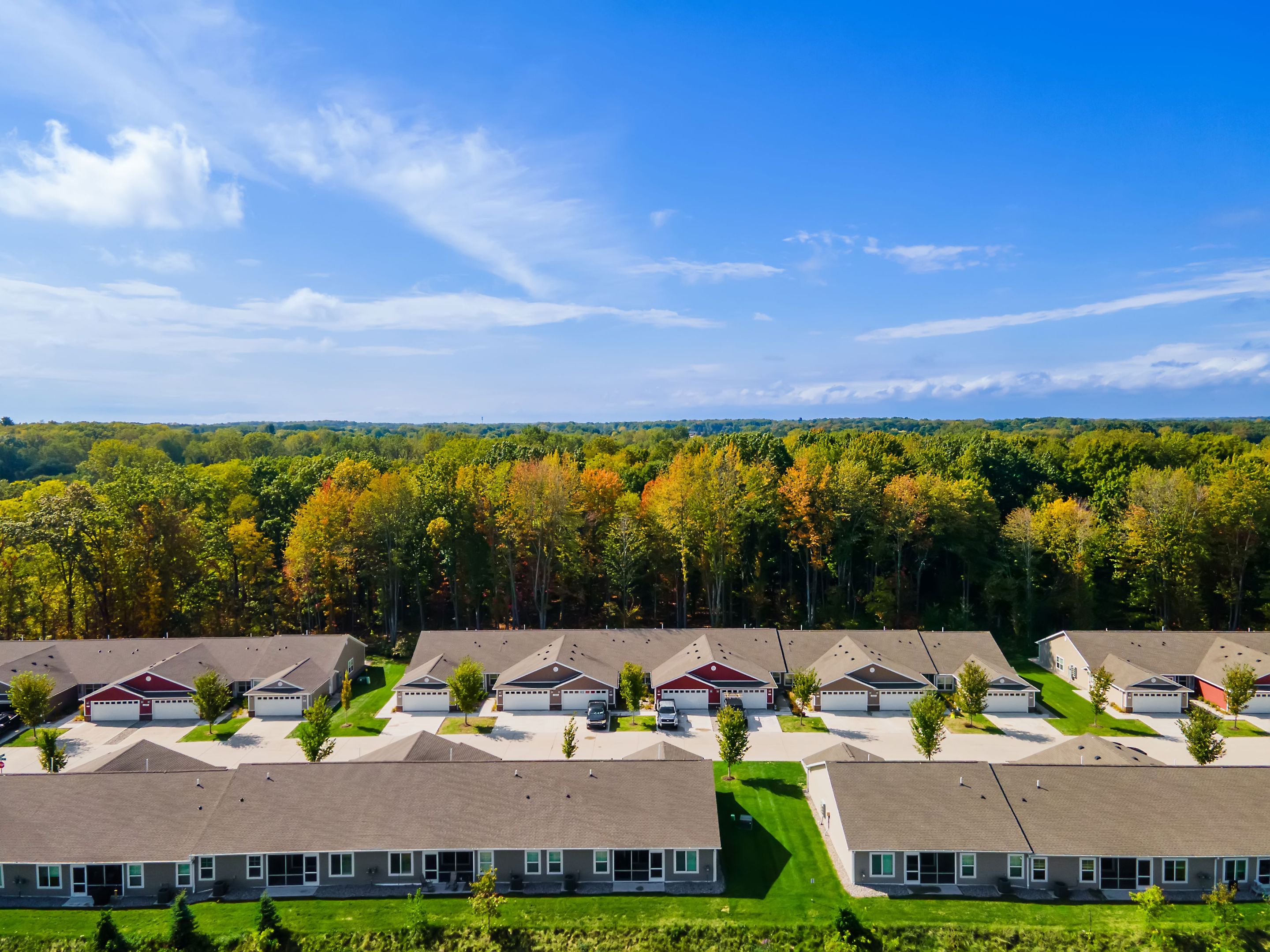 Aerial view of a suburban neighborhood with rows of single-story houses, green lawns, and a forested area in the background under a blue sky.