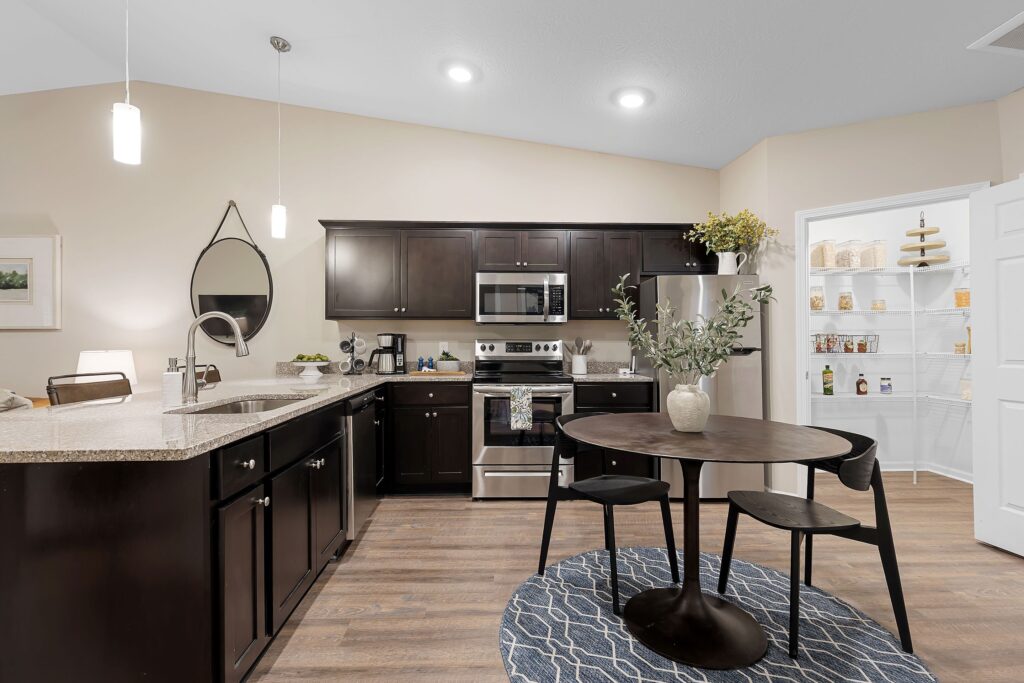 Modern kitchen with dark cabinets, stainless steel appliances, a small round dining table, and an open pantry stocked with food items.