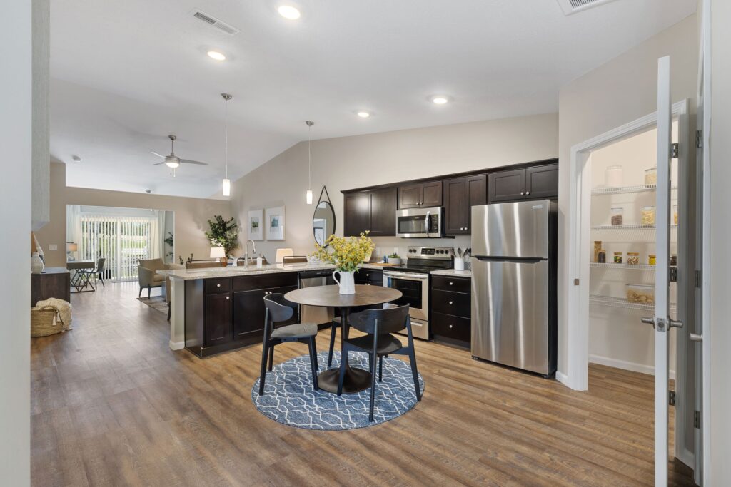 Modern kitchen with dark cabinets, stainless steel appliances, round dining table on blue rug, open pantry, and view into living and dining areas.
