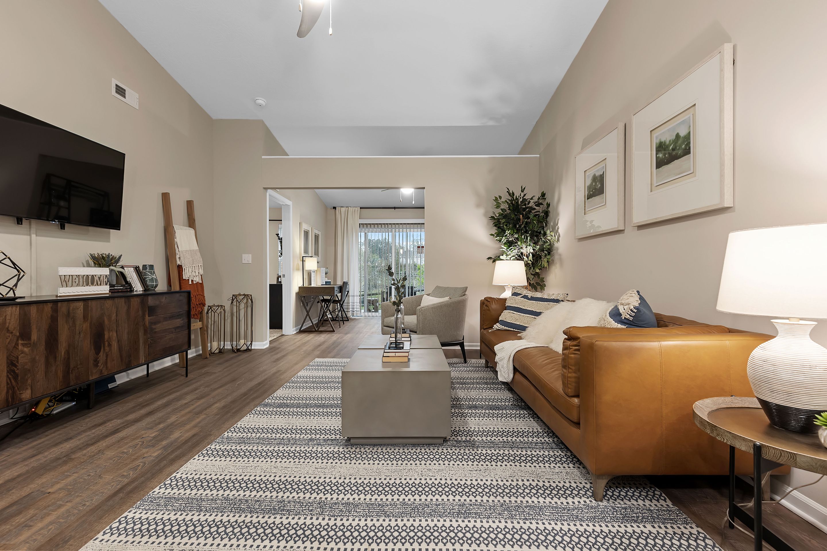 Modern living room with a brown leather sofa, patterned rug, wooden floor, wall art, and neutral decor; TV mounted on the wall and a coffee table in the center.