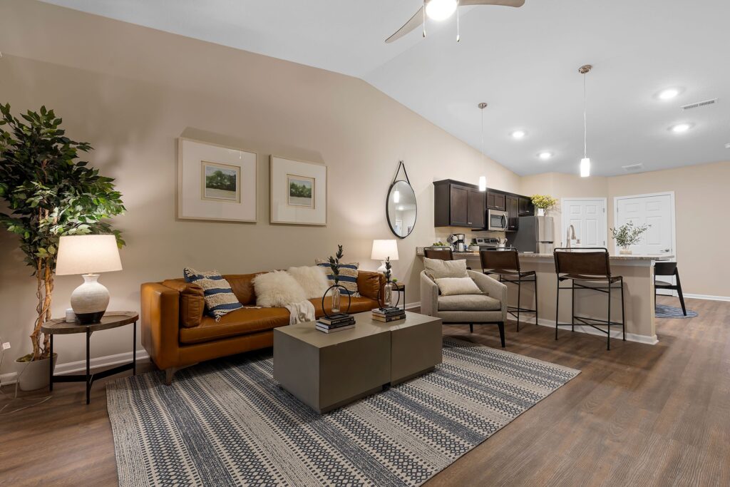 Open-concept living room and kitchen with brown sofa, striped rug, bar stools at counter, pendant lights, and neutral decor.