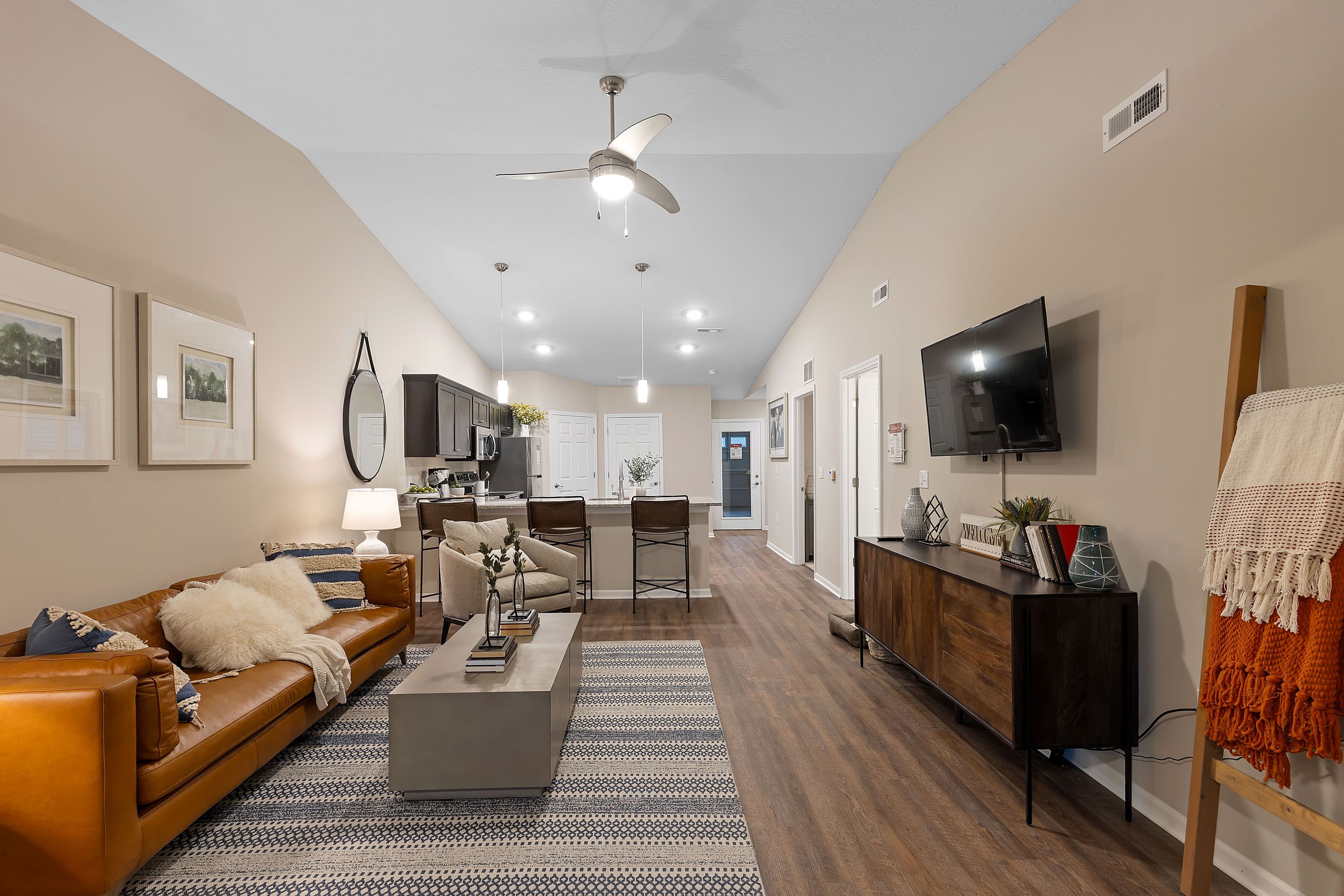 Open-concept living room and kitchen with a brown leather sofa, modern furniture, wall-mounted TV, island seating, light wood floors, and neutral-colored walls.