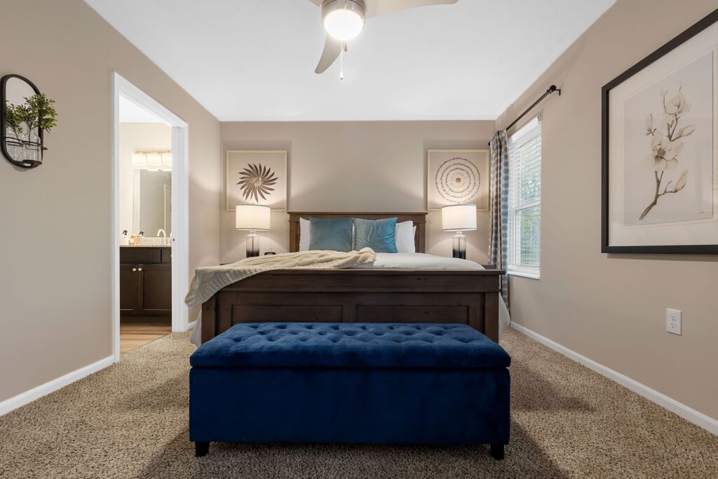 A neatly arranged bedroom features a dark wooden bed with blue pillows, two bedside lamps, a blue upholstered bench, carpeted floor, and artwork on the walls.
