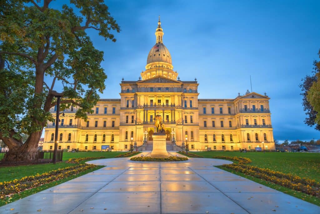 The Michigan State Capitol building in Lansing is illuminated at dusk, with a statue and trees in the foreground.