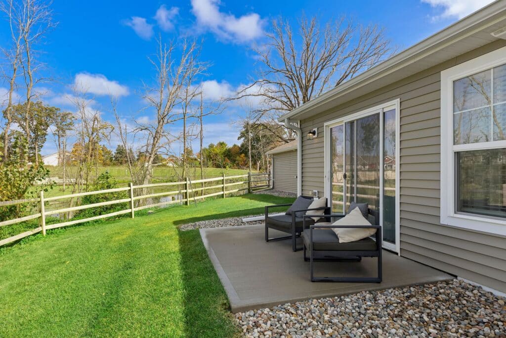 A small concrete patio with two chairs is adjacent to a beige house; the yard is fenced, with grass and trees under a blue sky with scattered clouds.