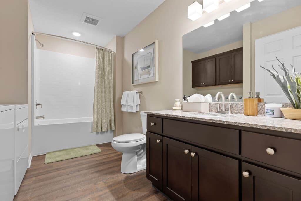 Modern bathroom with dark wood cabinets, granite countertop, white toilet, bathtub with shower curtain, wall art, and towel racks on light-colored walls.
