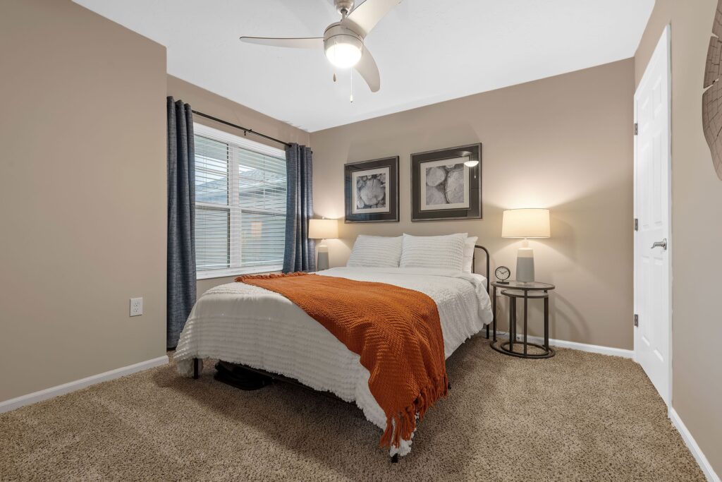 A neatly made bed with a white comforter and orange throw, two lamps on side tables, framed art above the headboard, carpeted floor, and a ceiling fan in a cozy bedroom.