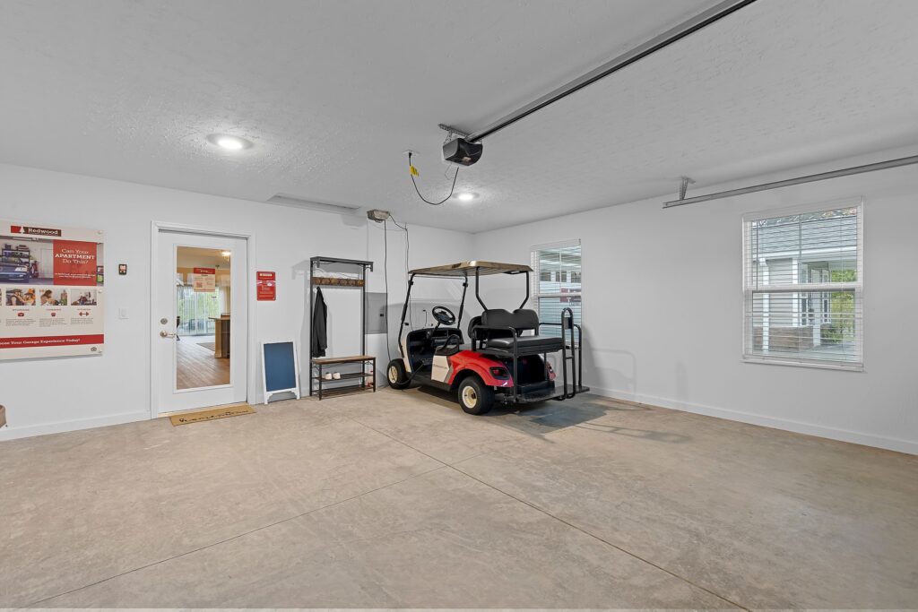 A clean, spacious garage with a red and black golf cart parked inside, white walls, large windows, a door, and informational posters on the wall.