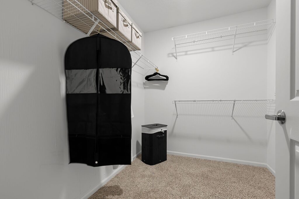 Empty walk-in closet with carpeted floor, wire shelving, hanging garment bag, black hanger, storage bins, and a black laundry basket.