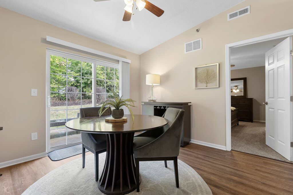 A small dining area with a round table, two chairs, a lamp, and a plant, set near sliding glass doors with a view of a backyard. An open door leads to a bedroom.