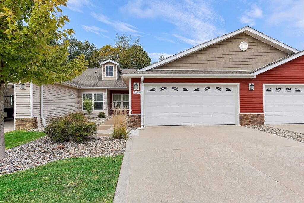 Single-story townhouse with a double garage, red and beige siding, small front porch, and landscaped yard with rocks and shrubs under a partly cloudy sky.