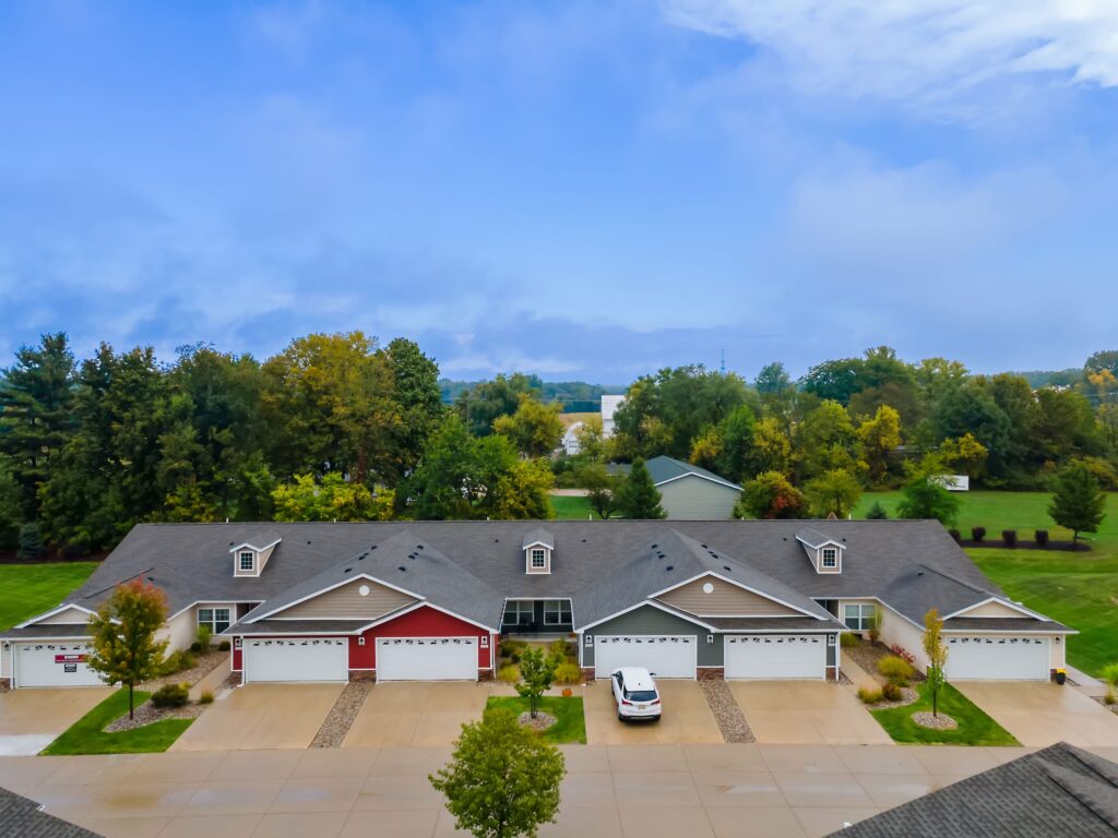 A row of attached townhouses with garages and driveways, a single parked car, trees, a large grassy area, and a partly cloudy sky in the background.