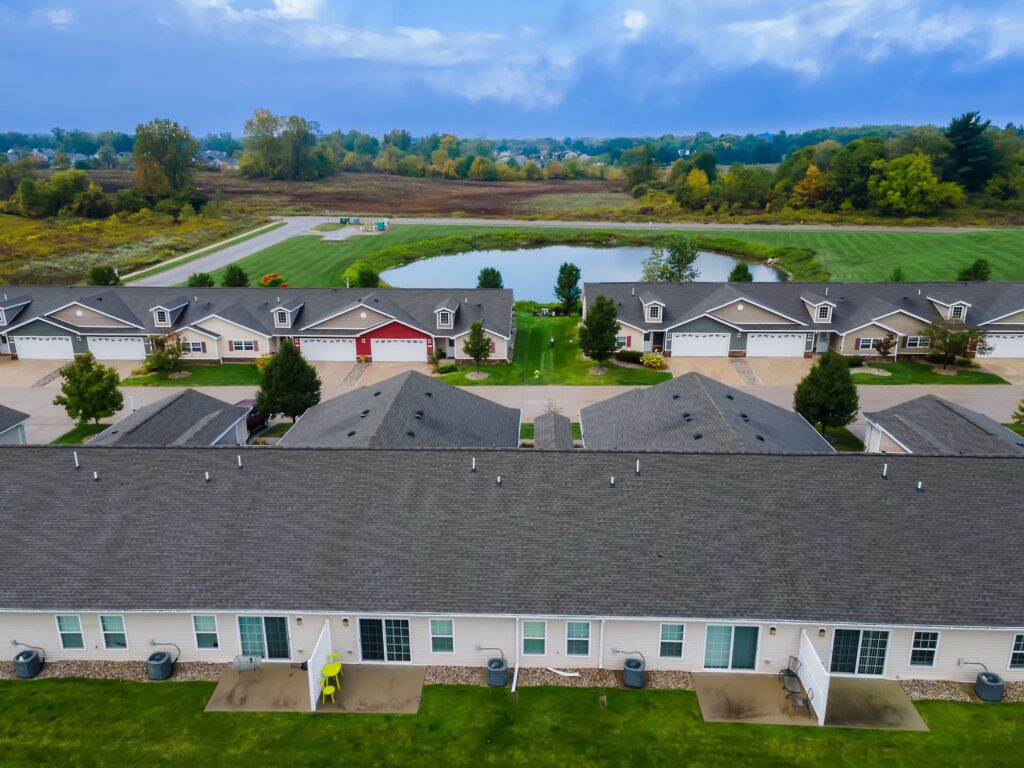 Aerial view of a suburban neighborhood with rows of houses, manicured lawns, a small pond, and trees in the background under a partly cloudy sky.