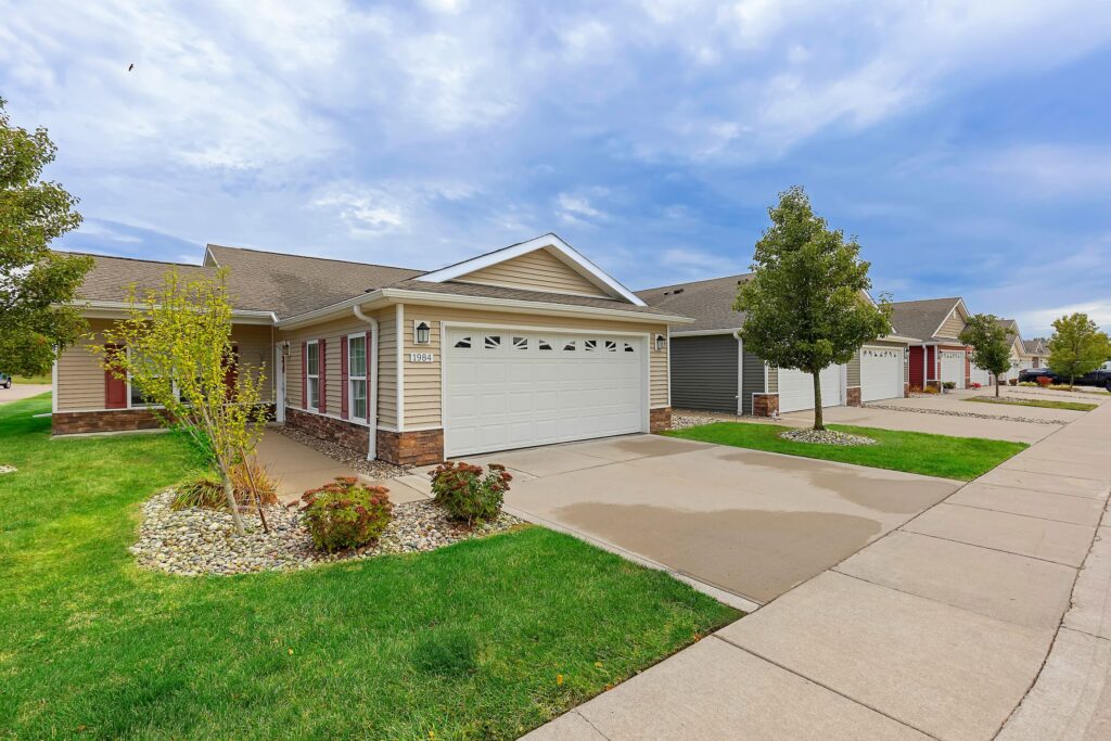 Single-story suburban house with a double garage, driveway, front lawn, and small trees along a quiet residential street under a partly cloudy sky.