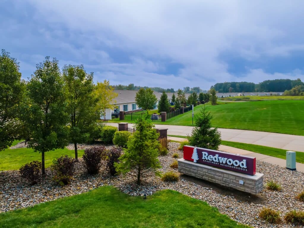 A landscaped entrance with a “Redwood Apartment Neighborhoods” sign, green grass, trees, shrubs, and a paved street under a cloudy sky.