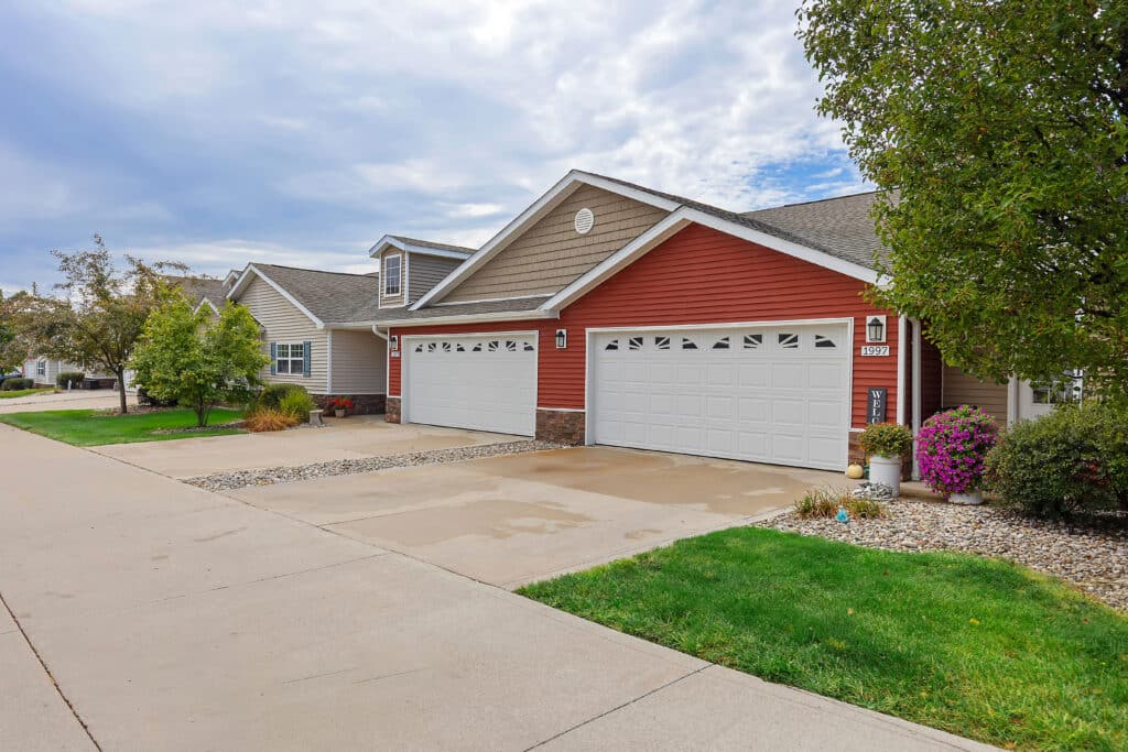 A row of suburban houses with attached three-car garages, landscaped lawns, and a wide concrete driveway under a partly cloudy sky.