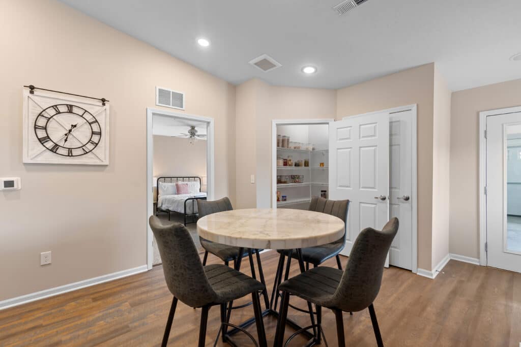 Round dining table with four chairs in a room with wood flooring; open doors reveal a bedroom and a pantry with shelves of food items. A large wall clock hangs on the left wall.