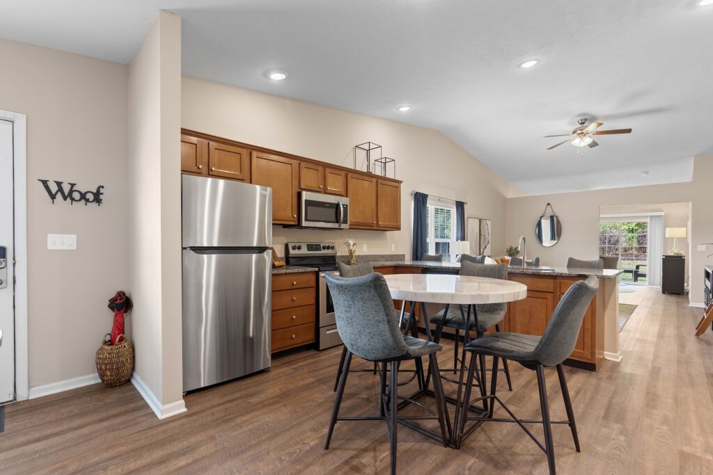 Open kitchen with stainless steel appliances, wooden cabinets, a round dining table with four chairs, and a decorative "Woof" sign on the wall.