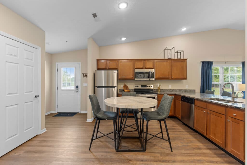 Modern kitchen with wooden cabinets, stainless steel appliances, a round dining table with four chairs, and a window letting in natural light.