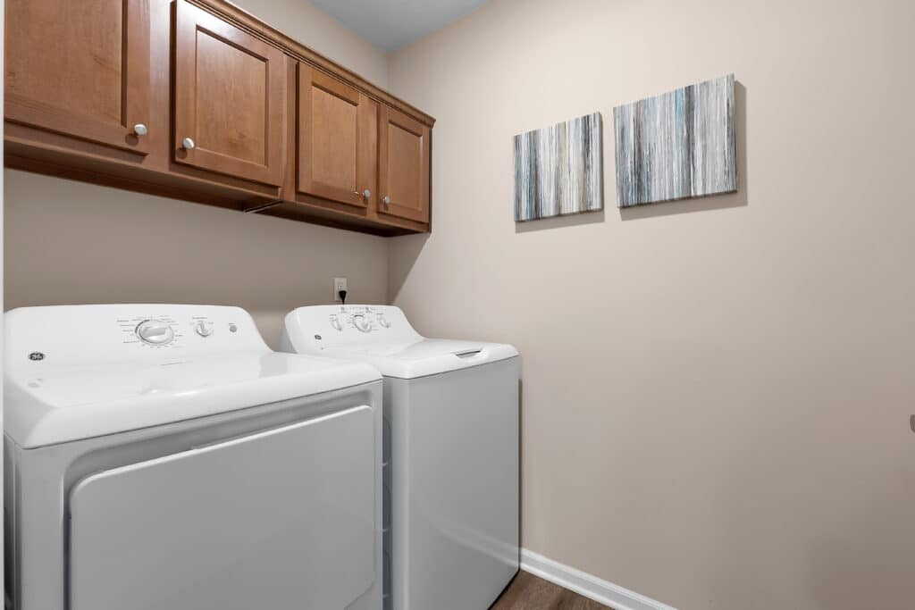 A laundry room with a white dryer and washer, wooden cabinets above, beige walls, and two abstract paintings hanging on the right side.