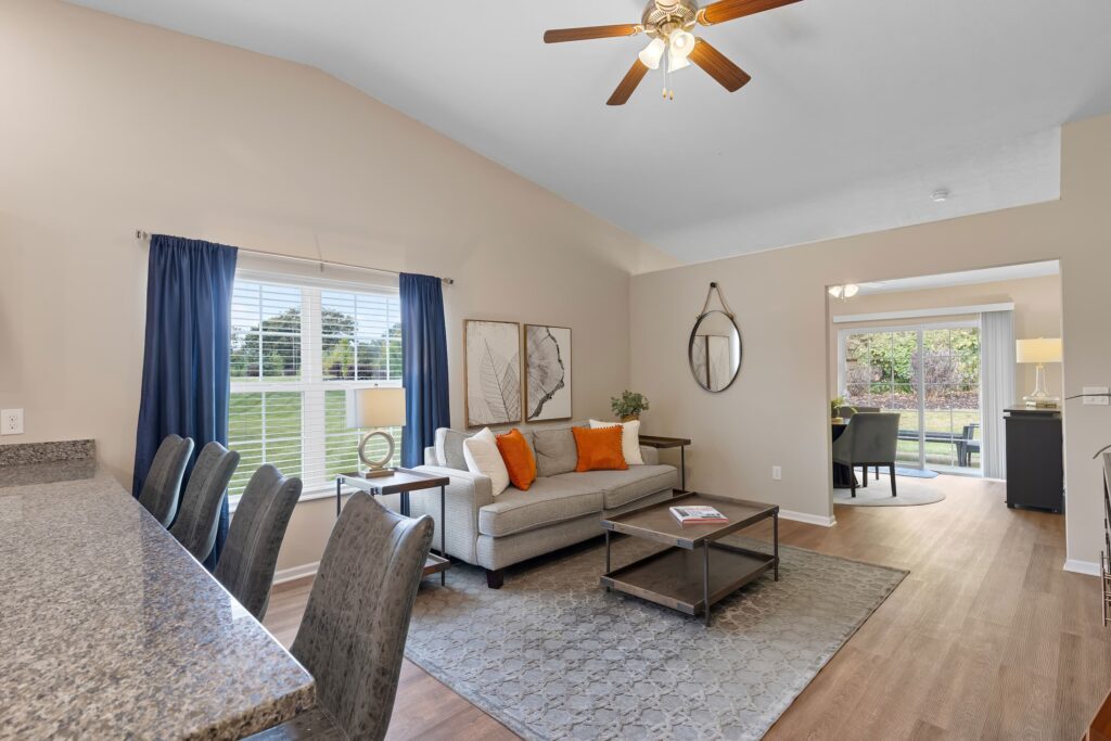 A living room with a sofa, coffee table, and chairs; large window with blue curtains, ceiling fan, and view into a sunlit adjoining room.