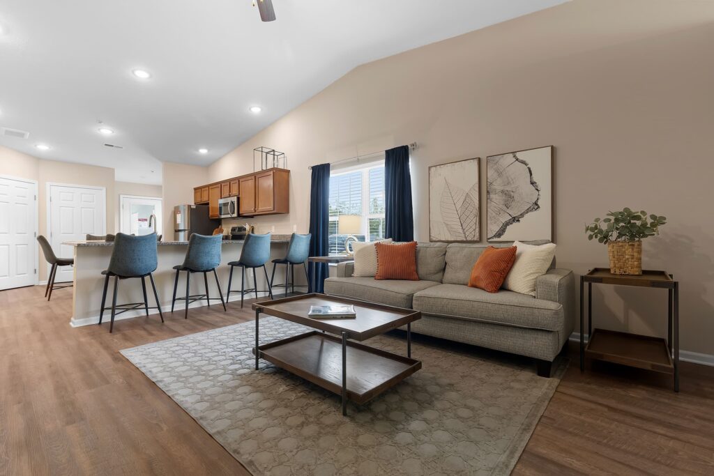 Open-concept living area with a gray sofa, blue bar stools at a kitchen island, wooden floors, and neutral decor with orange and blue accents.