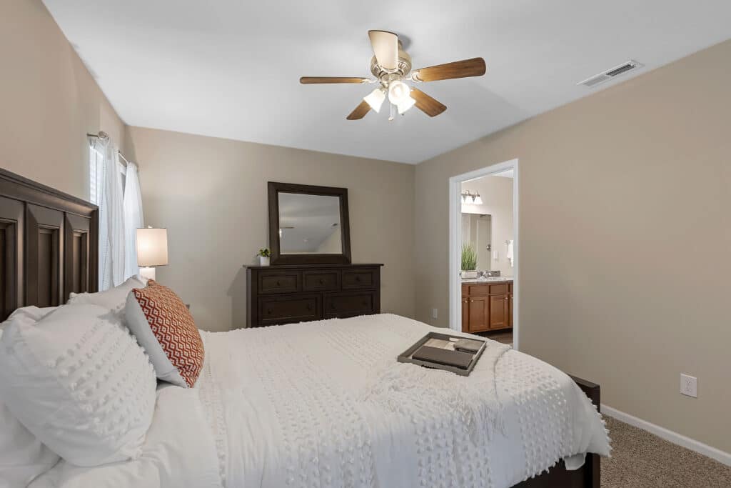A bedroom with a neatly made bed, a decorative pillow, a tray on the bed, a dresser with a mirror, and a ceiling fan, leading to a bathroom with dual sinks.