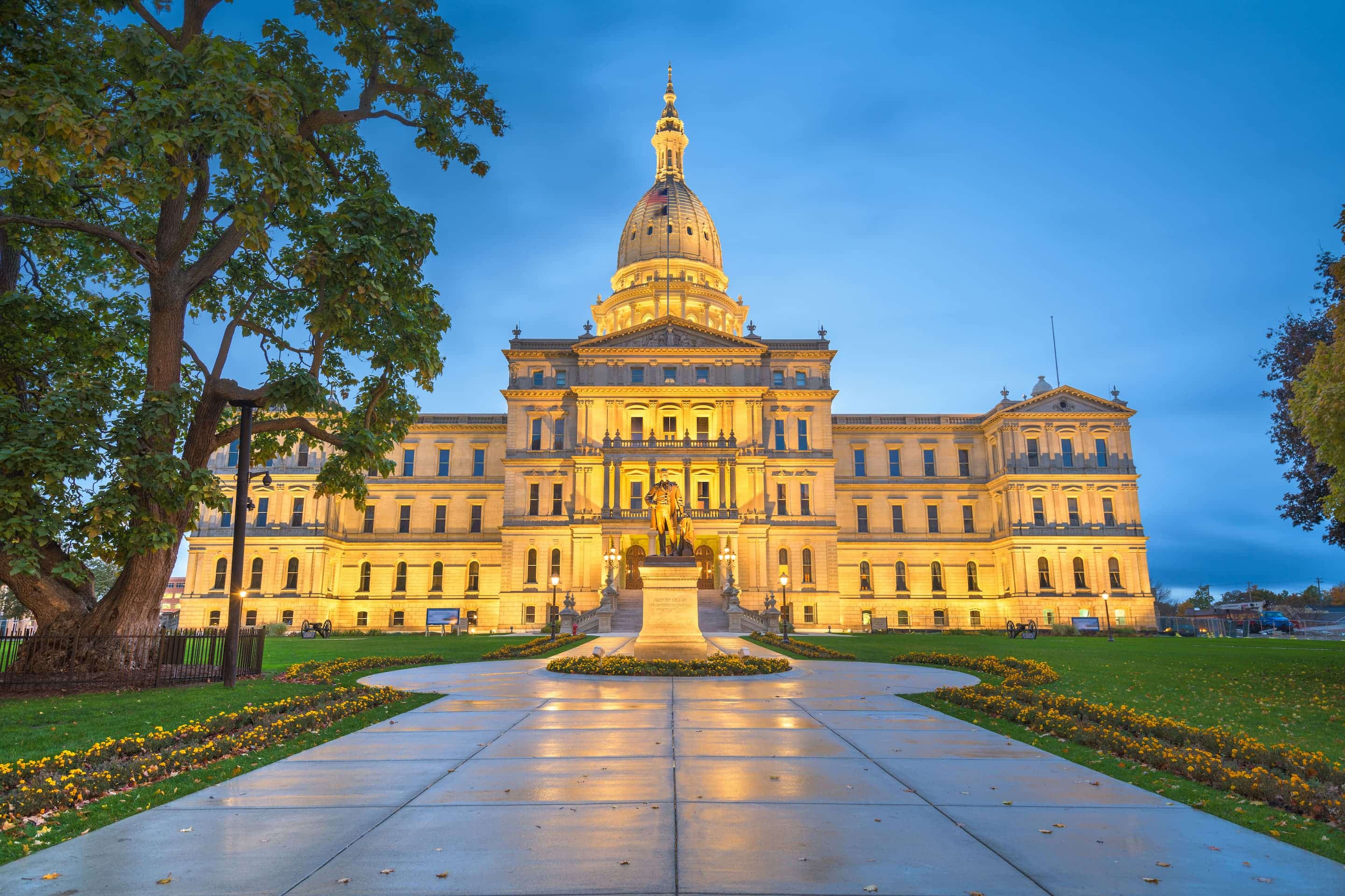 The Michigan State Capitol building in Lansing is illuminated at dusk, with a statue and pathway leading to the main entrance.