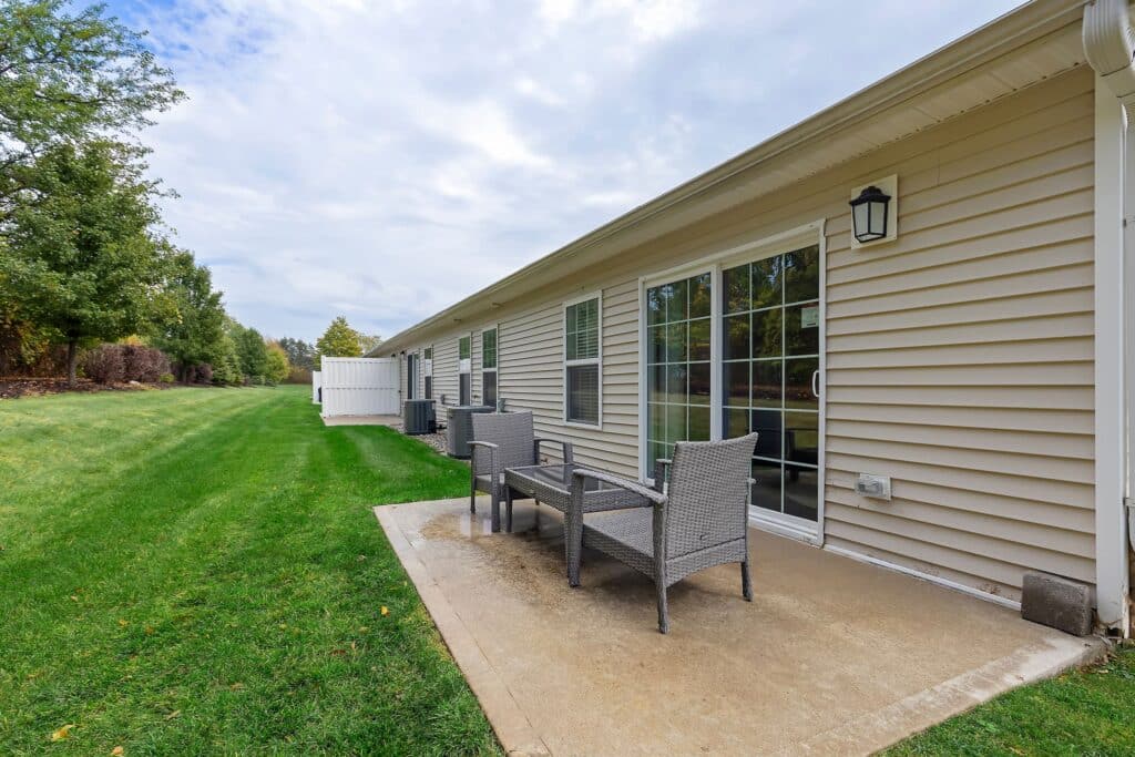 Concrete patio with wicker chairs and table outside a beige single-story building, adjacent to a grassy lawn under a partly cloudy sky.