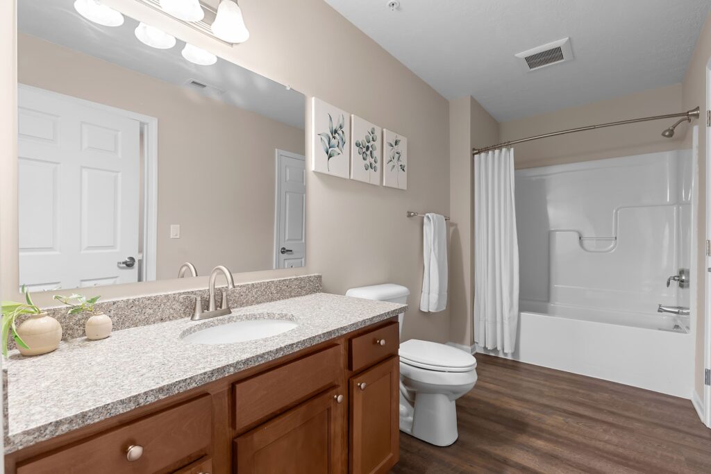 Modern bathroom with a granite countertop vanity, sink, toilet, towel rack, and a white shower-tub combo, featuring light walls and wood-style flooring.