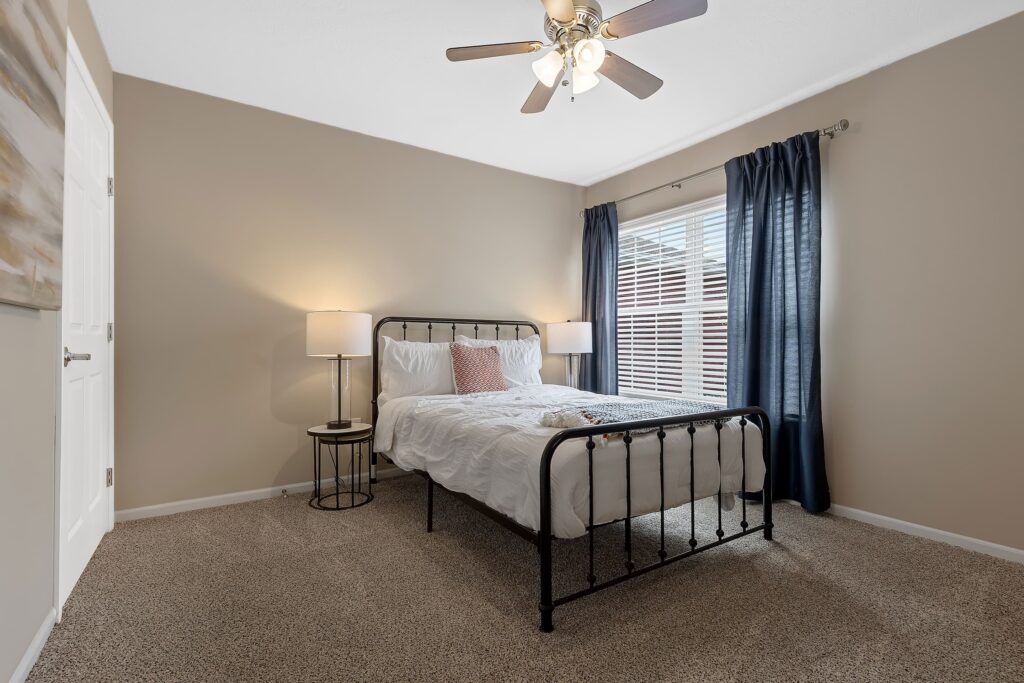 A simple bedroom with a black metal bed, white bedding, two side tables with lamps, beige walls, carpeted floor, ceiling fan, and a window with dark curtains.