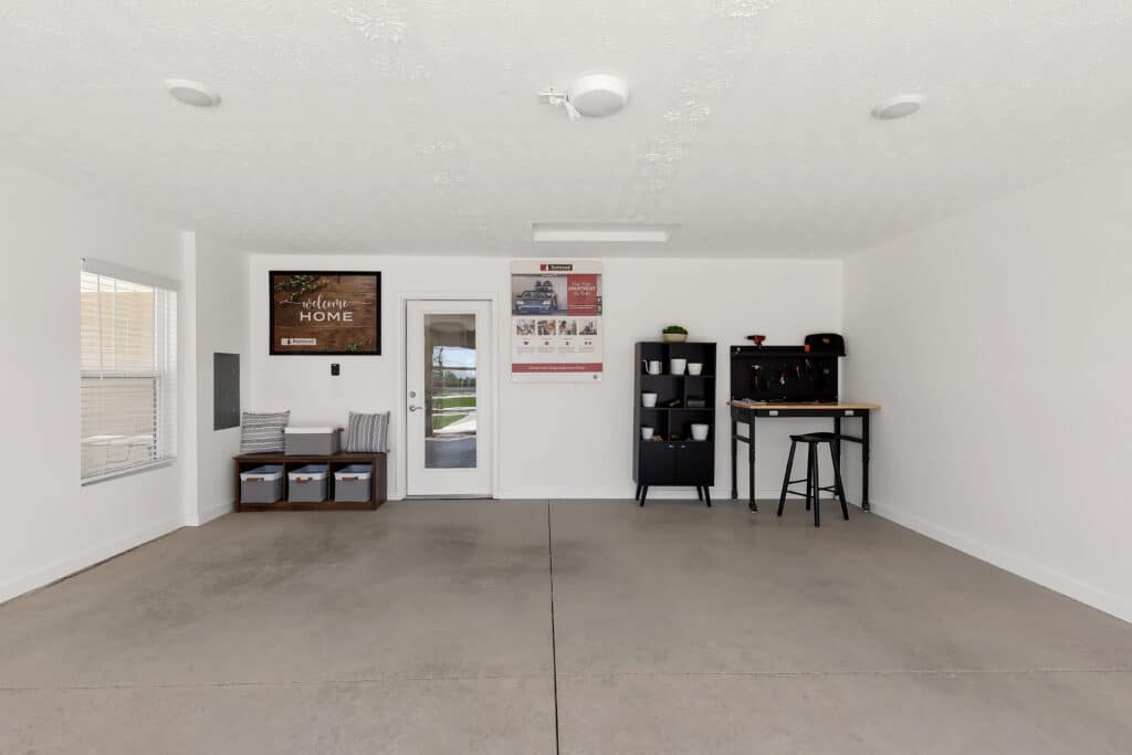 A clean, organized garage with a small sitting area, storage baskets, wall art, a shelving unit, and a high table with two stools against a white wall.