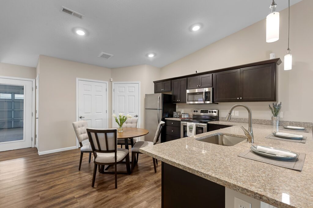 Modern kitchen with granite countertops, dark cabinets, stainless steel appliances, a round dining table with four chairs, and wood flooring under recessed and pendant lighting.