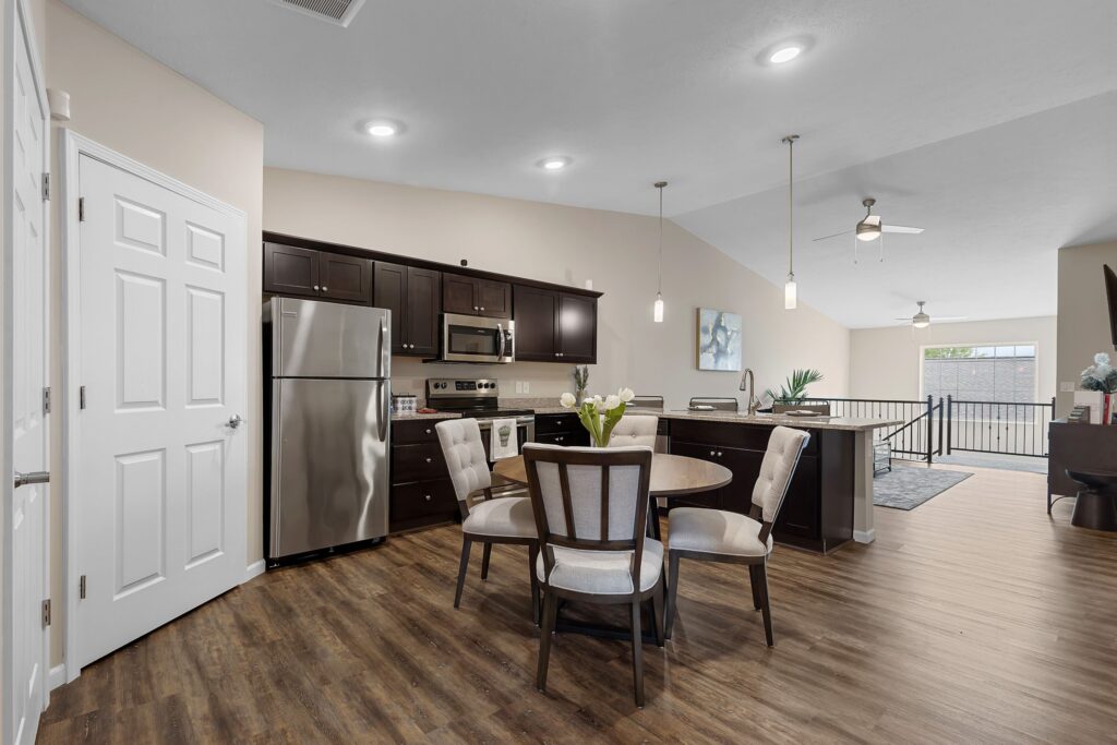 Modern kitchen with dark cabinets, stainless steel appliances, a round dining table with four chairs, wood flooring, and an open layout leading to a living area.