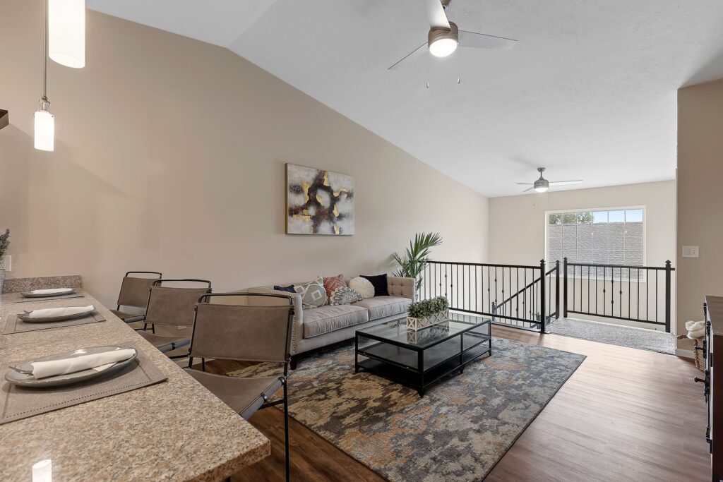 Open living area with a sofa, glass coffee table, chairs, and a rug; kitchen counter with place settings in the foreground; large window and ceiling fans visible.