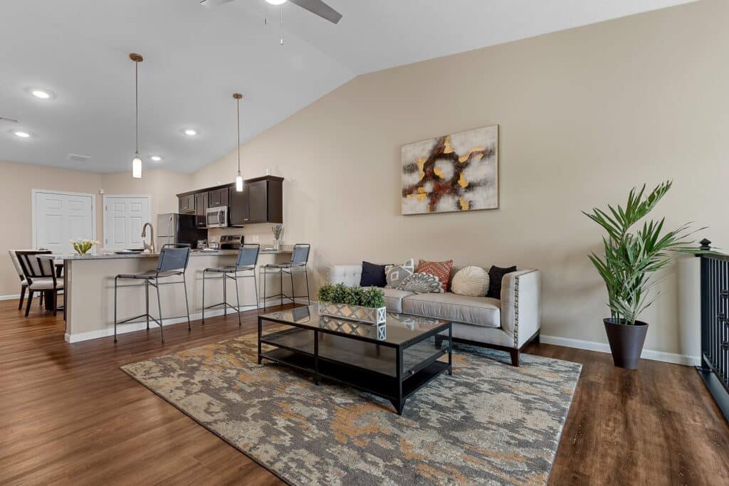 Modern open-plan living room and kitchen with a beige sofa, decorative pillows, coffee table, bar stools, plants, and abstract wall art on light walls with wood flooring.