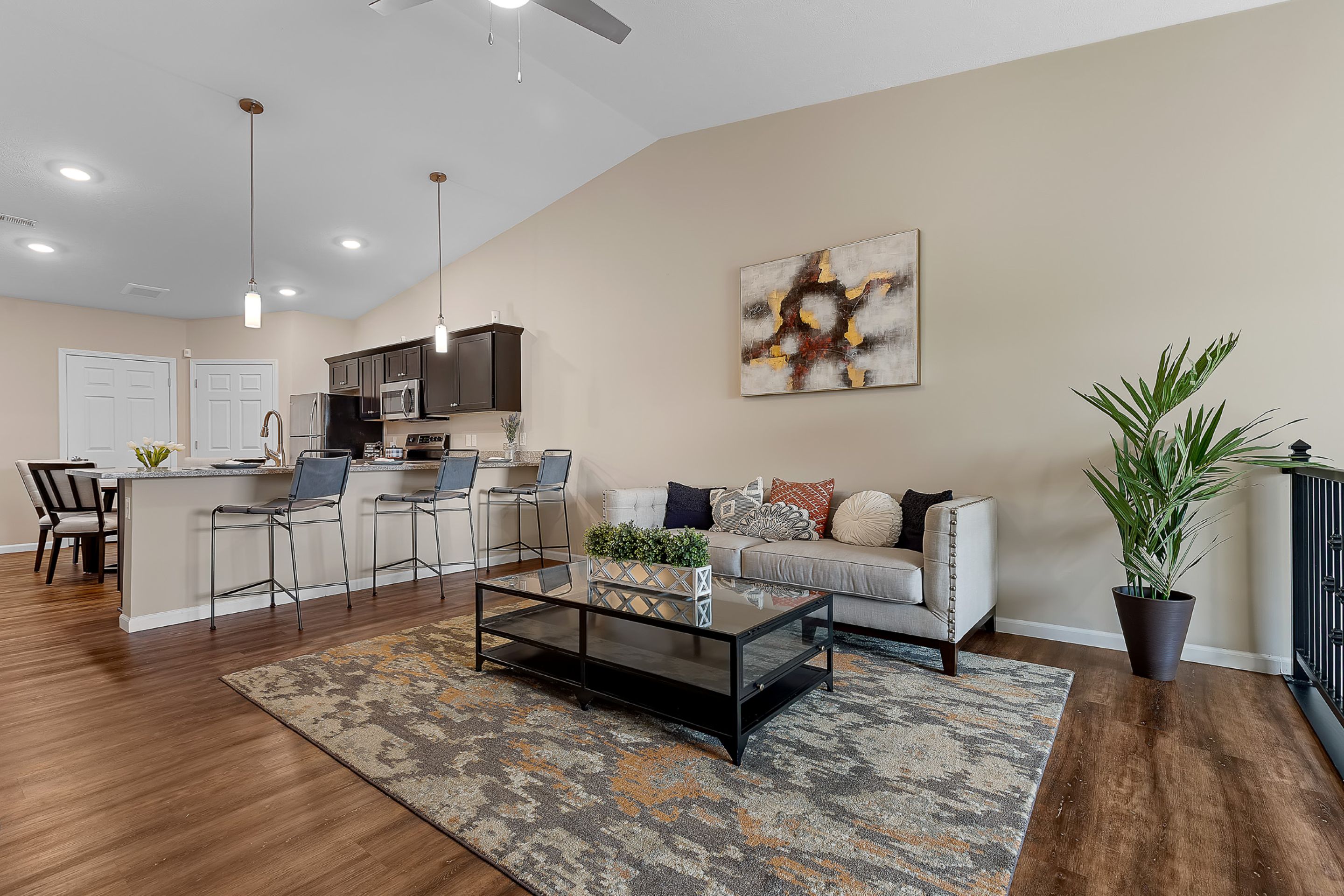 Modern open-plan living room and kitchen with a beige sofa, decorative pillows, coffee table, bar stools, plants, and abstract wall art on light walls with wood flooring.