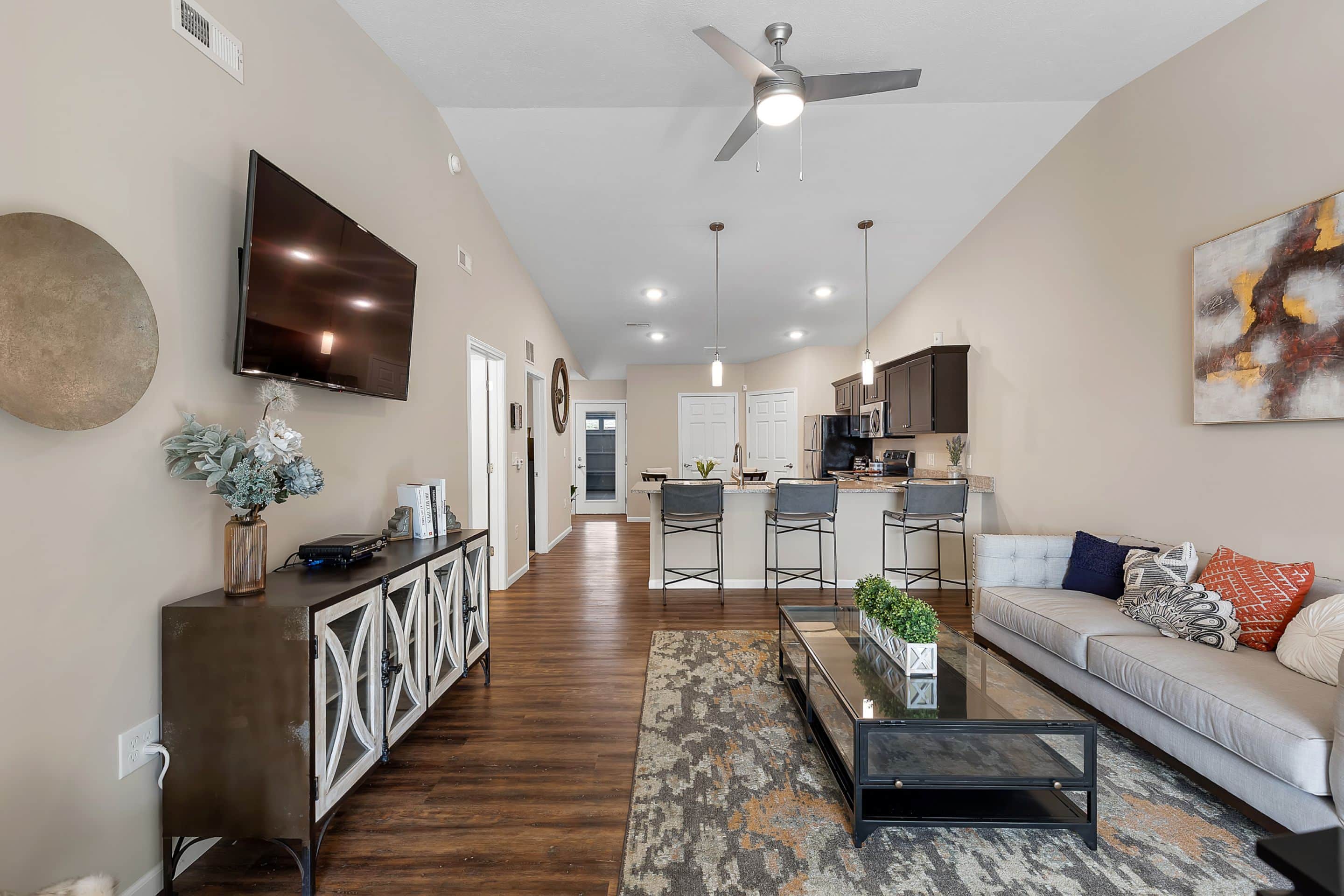 Modern open-concept living room and kitchen with neutral tones, ceiling fans, wall-mounted TV, sofa, bar stools, and decorative accents.