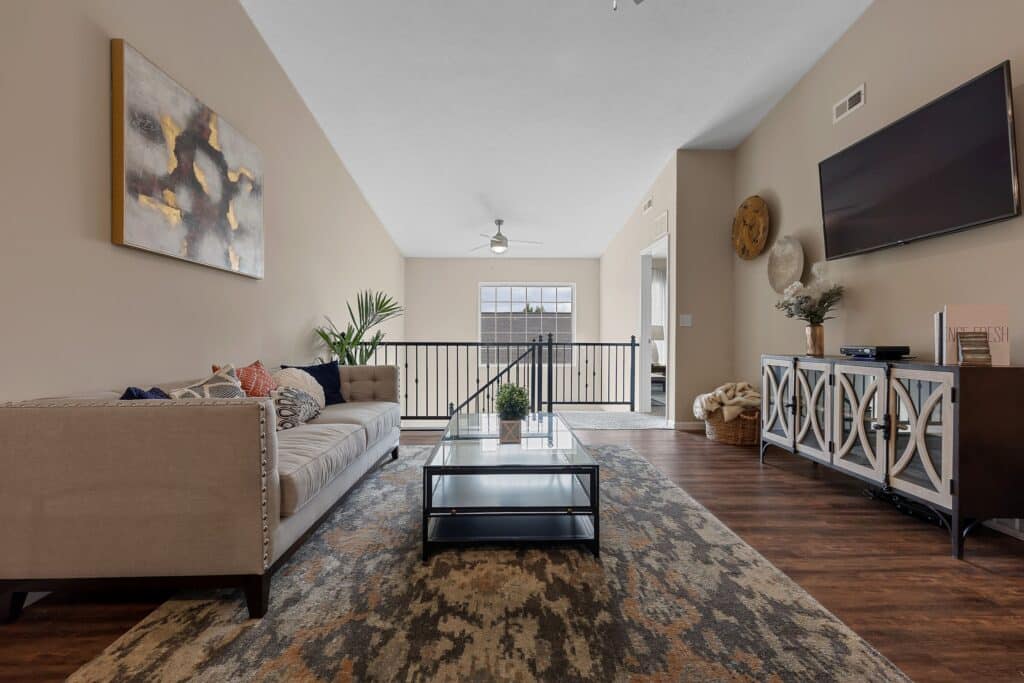 A modern living room with a beige sofa, glass coffee table, patterned rug, wall-mounted TV, and decorative console under natural light.